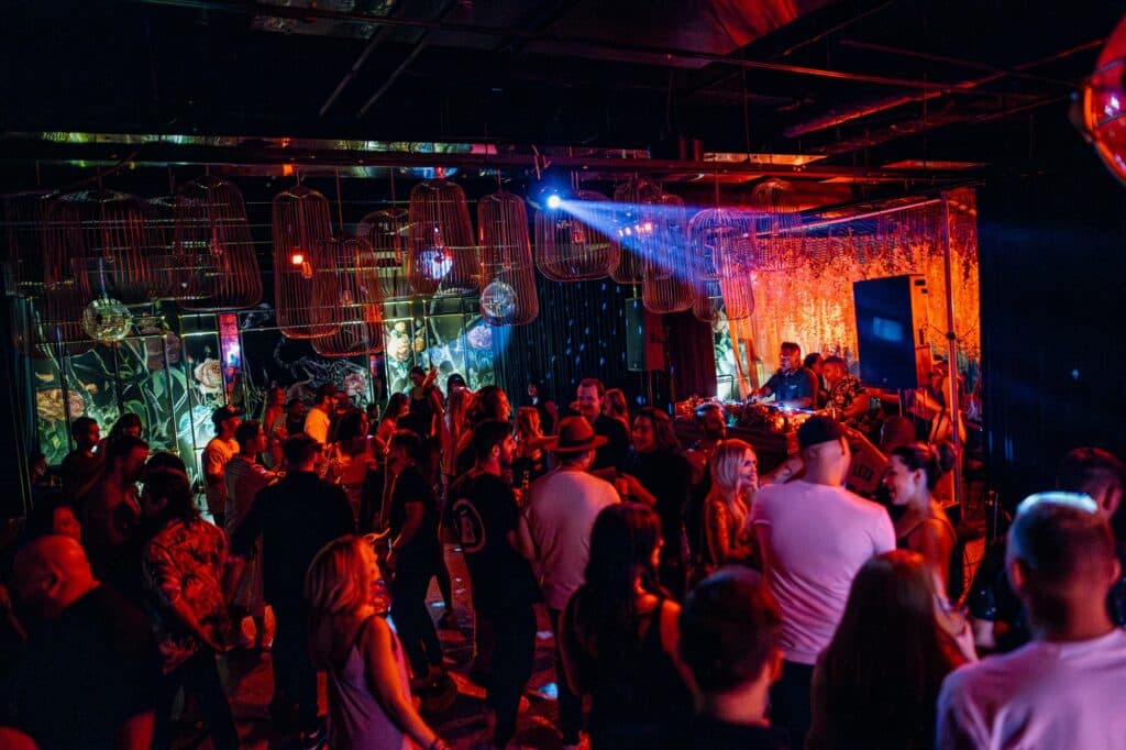 Crowded nightclub scene with people dancing under colorful lights and decorative hanging birdcage lanterns.