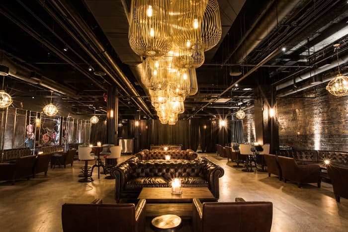 Moody industrial lounge with leather tufted sofas and a row of golden cage pendant lights.
