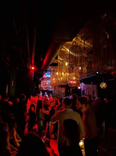 Crowded nightclub with people dancing under large birdcage chandeliers and atmospheric red and yellow lighting.