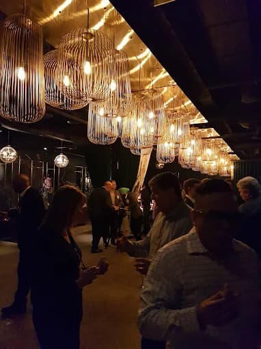 People socialize at a dimly lit event under large, warm, decorative cage-style pendant lights.